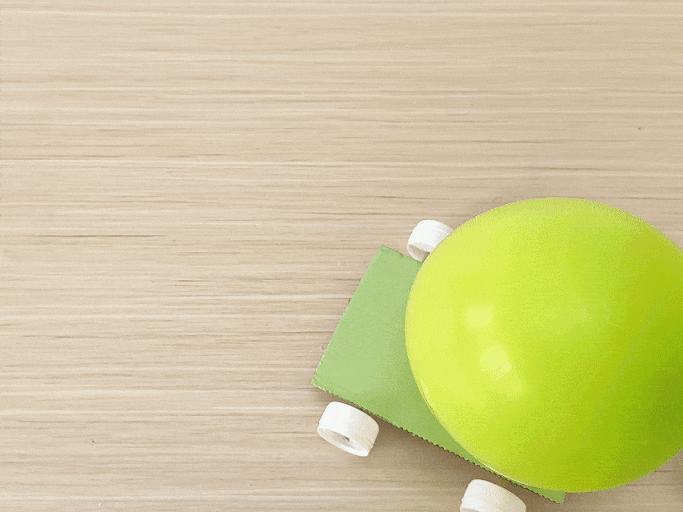 A small, homemade balloon-powered car moves across a light wooden surface. The car is constructed from a piece of light green cardboard, with white bottle caps serving as wheels. A lime green inflated balloon is attached to the cardboard. As the air is released from the balloon, it propels the car forward, demonstrating a simple physics principle. The camera follows the car from a top-down perspective as it travels across the wooden surface.