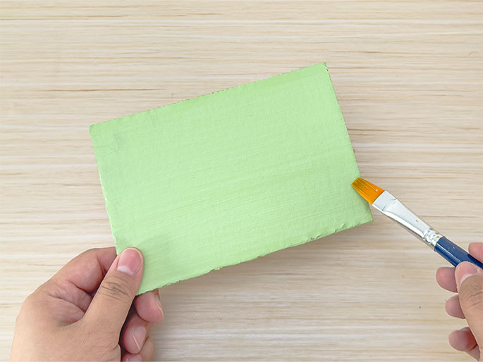 Two hands are shown in the process of painting a piece of cardboard light green. One hand holds the cardboard, which is mostly covered in the light green paint, against a light woodgrain background. The other hand holds a paintbrush with orange bristles and a blue handle, applying more paint to the corner of the cardboard.