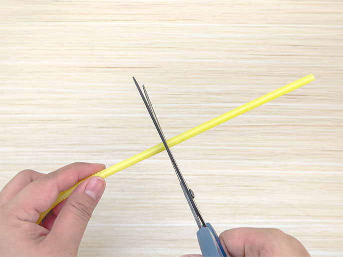 Two hands are shown cutting a yellow flexible drinking straw with a pair of scissors. One hand holds the straw against a light woodgrain background, while the other hand operates the gray-handled scissors, cutting the straw. The focus is on the action of cutting the straw.