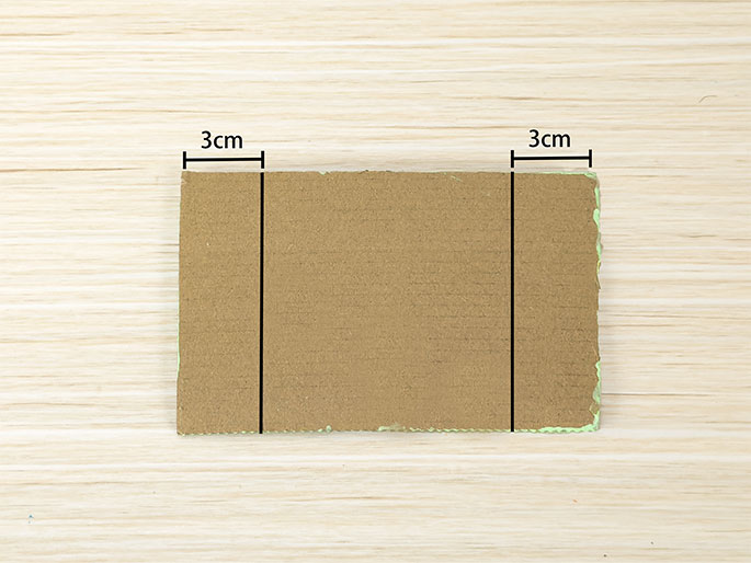 A rectangular piece of light brown cardboard rests on a light woodgrain surface. Two vertical black lines divide the cardboard into three equal sections. Black lines and text indicate that the outer sections are each 3cm wide. This image illustrates measurements marked on the cardboard, likely for cutting. A faint trace of light green paint is visible along the bottom edge of the cardboard.