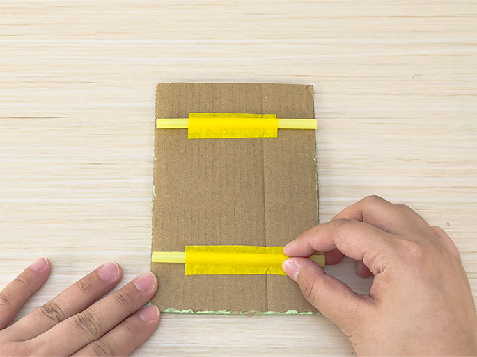 Two hands are shown attaching yellow straws to a piece of cardboard. The cardboard, placed on a light woodgrain background, has two short yellow straws horizontally positioned, one near the top and the other near the bottom. Bright yellow tape secures the straws to the cardboard. One hand holds the cardboard steady, while the other hand adjusts the tape around one of the straws. A trace of light green paint is visible along the bottom edge of the cardboard.