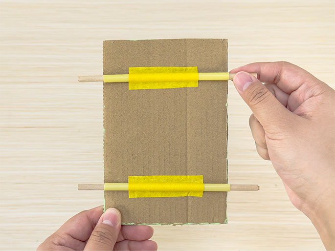 Two hands hold a piece of light brown cardboard against a light woodgrain background. Two thin wooden dowels are attached horizontally to the cardboard, one near the top and the other near the bottom. Sections of yellow straws are taped to the center of each dowel with bright yellow tape. The hands hold the dowels, extending beyond the edges of the cardboard. A faint trace of light green paint is visible along the bottom edge of the cardboard.