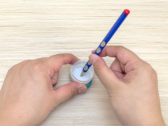 Two hands manipulate a pencil and a small plastic bottle cap on a light woodgrain surface. One hand holds the teal-colored base of the bottle cap, which has a white interior and lid. The other hand holds a blue pencil with a red eraser, positioning the pencil's point in the center of the bottle cap's white lid. The image suggests the pencil is being used to mark or pierce the center of the cap.