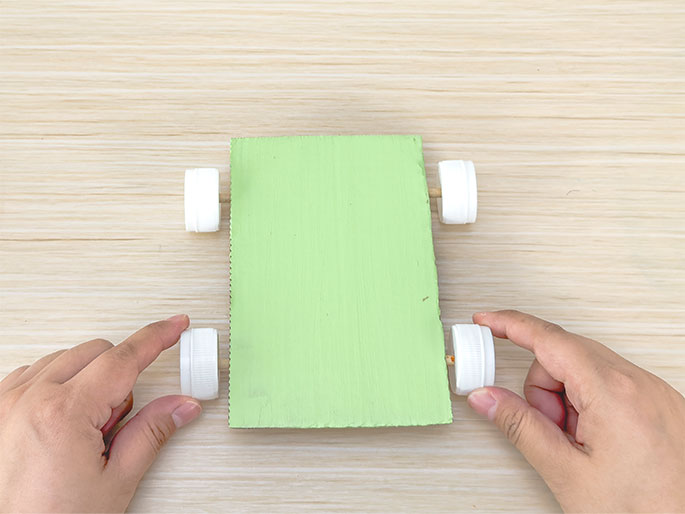 Two hands display a simple toy car constructed from a piece of light green painted cardboard and four white plastic bottle caps as wheels. The car rests on a light woodgrain surface. Short wooden dowels, acting as axles, connect the bottle cap wheels to the cardboard body. The hands hold the car from the sides, gently touching two of the wheels.