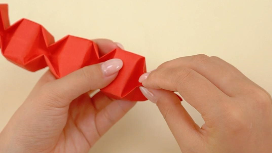 Fingers carefully manipulate and fold a section of a red origami creation, which appears to be a segmented or linked structure.