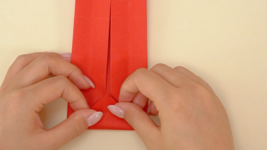 Two hands delicately fold the bottom edge of a red piece of paper, likely origami, to create a point or arrow shape.