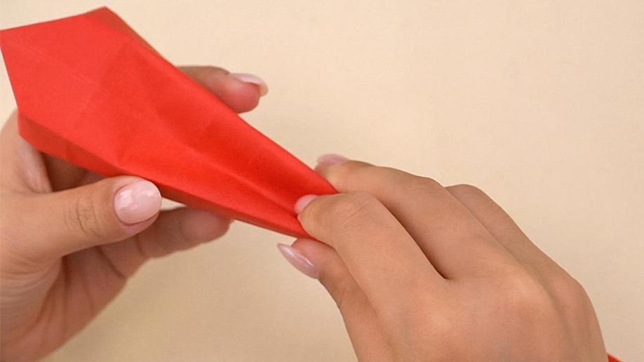 Hands pinch and shape a folded piece of red paper, appearing to create a more three-dimensional form, possibly a beak or nose-like shape, as part of an origami creation.