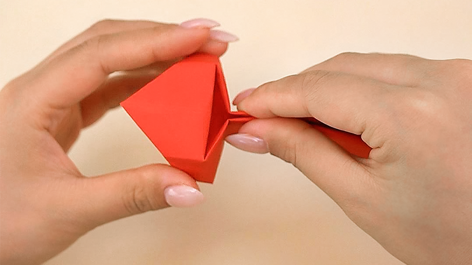 Two hands manipulate a folded, triangular piece of red origami paper, gently pulling a flap or section outwards.