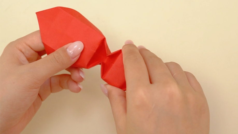 Two hands fold and shape a piece of red origami paper, creating a small, three-dimensional form with a defined crease or bend in the middle.