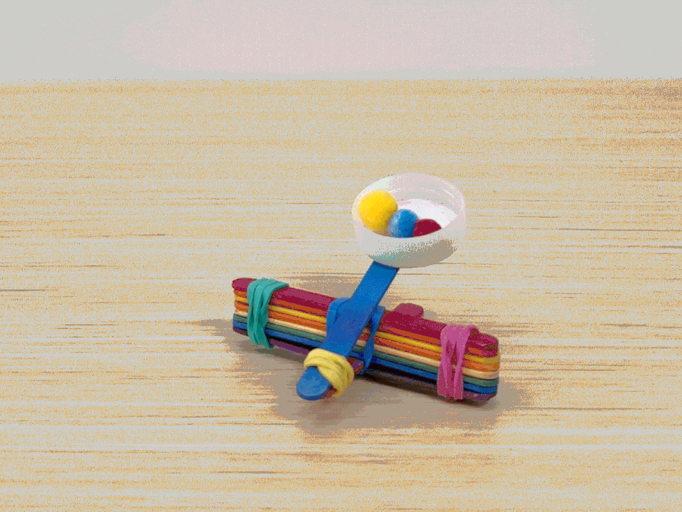 A small catapult made of colorful popsicle sticks, rubber bands, and a bottle cap launches pompoms across a wooden table. Hands are visible preparing the catapult for launch.