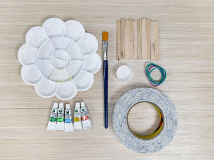 An overhead flatlay image displays various craft supplies arranged on a light wood-grain surface. The supplies include a flower-shaped paint palette, a paintbrush, small tubes of paint in various colors, a bundle of wooden craft sticks, a small container of white paint or glue, a collection of colorful rubber bands, and a roll of double-sided tape. These items suggest preparation for a children's art or craft project.