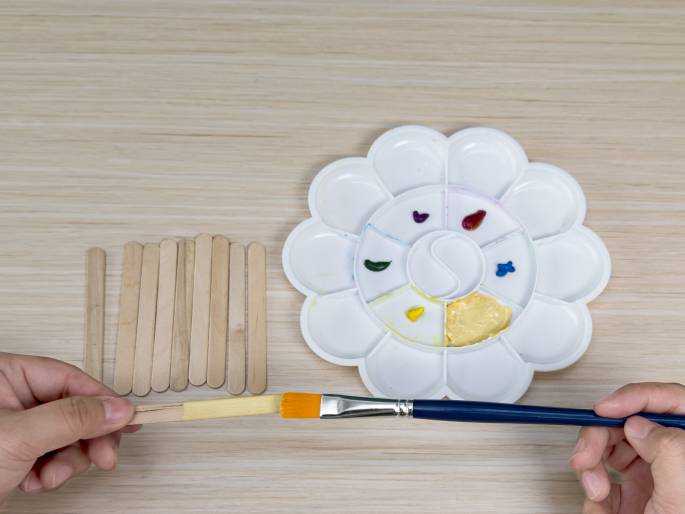 Two hands are painting a wooden craft stick yellow on a light wood-grain surface. A flower-shaped paint palette with dabs of various colored paints sits nearby. A bundle of unpainted craft sticks lies next to the palette. This image appears to capture a step in a crafting activity.