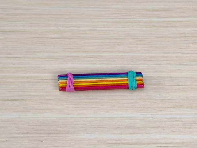 A bundle of colorful craft sticks lies on a light wood-grain surface. The sticks are arranged to create a rainbow effect, and they are held together by rubber bands at each end. One rubber band is pink, and the other is teal. This image likely depicts a finished craft project or a step in a larger project.