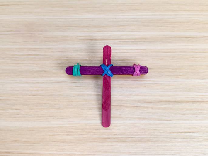 A simple cross made of popsicle sticks rests on a light wood-grain surface. The cross is constructed from two magenta-colored sticks, fastened together with small rubber bands. A teal rubber band holds the horizontal stick on the left, a doubled-over purple rubber band secures the center, and a pink rubber band holds the horizontal stick on the right. This image likely depicts a children's craft project.