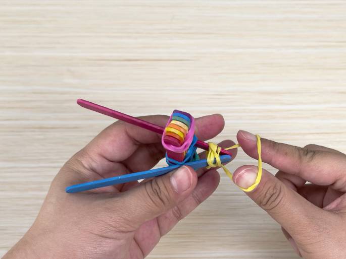 Two hands work on constructing a small projectile launcher or similar device made of popsicle sticks and rubber bands against a light wood-grain backdrop. One hand holds the main body of the device, composed of a stack of rainbow-colored sticks secured with pink rubber bands and two blue sticks forming a handle-like structure. The other hand stretches a yellow rubber band, likely to power the launcher's mechanism. A magenta stick appears positioned as part of the launching mechanism. The image captures a mid-construction step in a craft project.