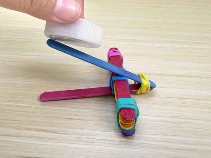 A hand positions a white plastic bottle cap onto a small catapult or launcher crafted from popsicle sticks and rubber bands on a light wood-grain surface. The launcher's base is constructed from a stack of rainbow-colored sticks bound together with rubber bands, and the launching arm appears to be a blue popsicle stick. Magenta-colored sticks and additional rubber bands form the support structure. The image likely demonstrates the final step in assembling the popsicle stick launcher, with the bottle cap serving as the projectile.