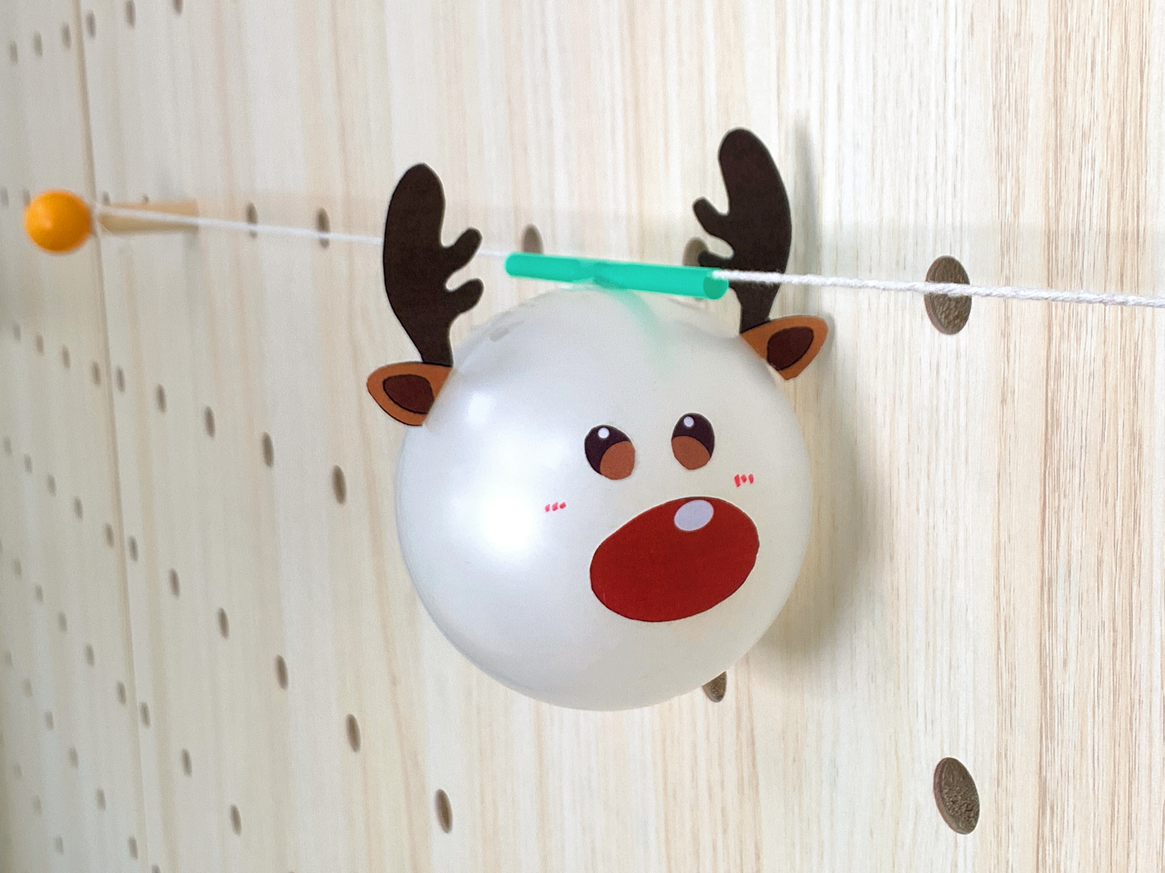 A reindeer-faced balloon, crafted with paper antlers and facial features, is attached to a short green straw threaded onto a string strung across a wooden pegboard.