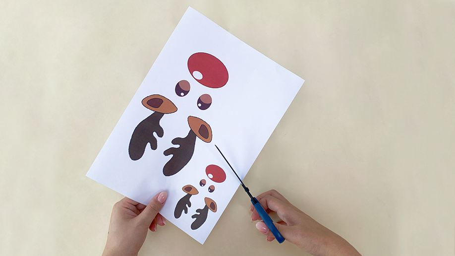 A pair of hands use scissors to cut out reindeer facial features from a printed sheet of paper.