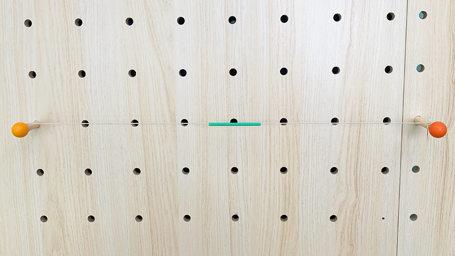 A piece of string is stretched taut between two orange-tipped wooden pegs on a pegboard, with a small green piece of straw or tube threaded onto the string.