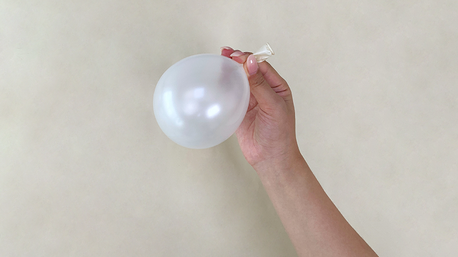 A hand holds a small, partially inflated, translucent white balloon against a plain, cream-coloured background.