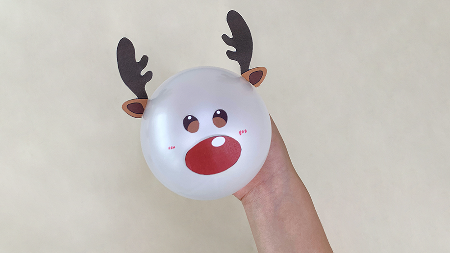 A hand holds a white balloon decorated with a reindeer face, complete with a red nose, cute eyes, rosy cheeks, and dark brown antlers.