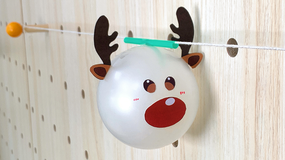 A reindeer-faced balloon, crafted with paper antlers and facial features, is attached to a short green straw threaded onto a string strung across a wooden pegboard.