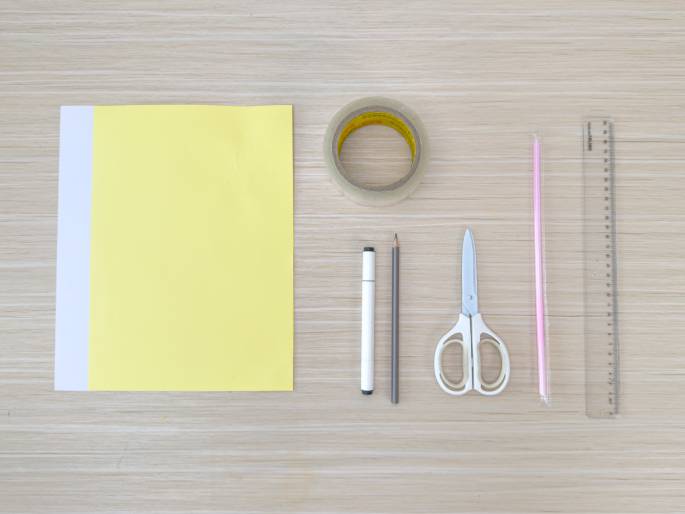 An overhead flatlay image displays the materials for a craft project arranged on a light wood-grained surface. The materials include a sheet of pale yellow paper partially overlaid with a smaller sheet of white paper, a roll of clear tape, a white marker, a pencil, a pair of white-handled scissors, a pink straw, and a clear plastic ruler. These items are neatly laid out, suggesting preparation for a paper or DIY project.