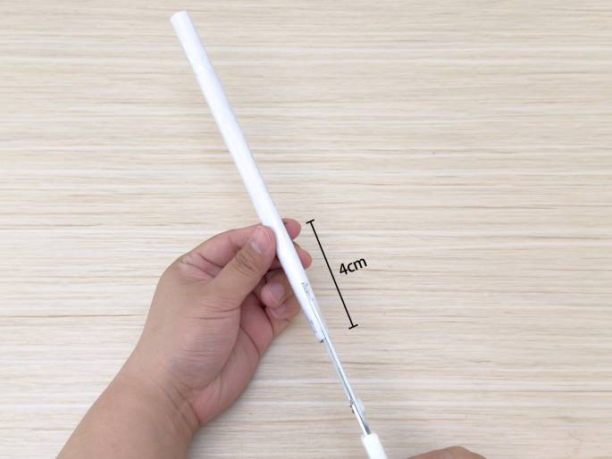Hands are using white-handled scissors to cut a white drinking straw against a light wood background. A black vertical line indicates a measurement of 4cm, showing the approximate length of the portion of the straw being cut. The left hand holds the straw, and the right hand operates the scissors. The image likely illustrates a step in a craft project where precise measurements are important.