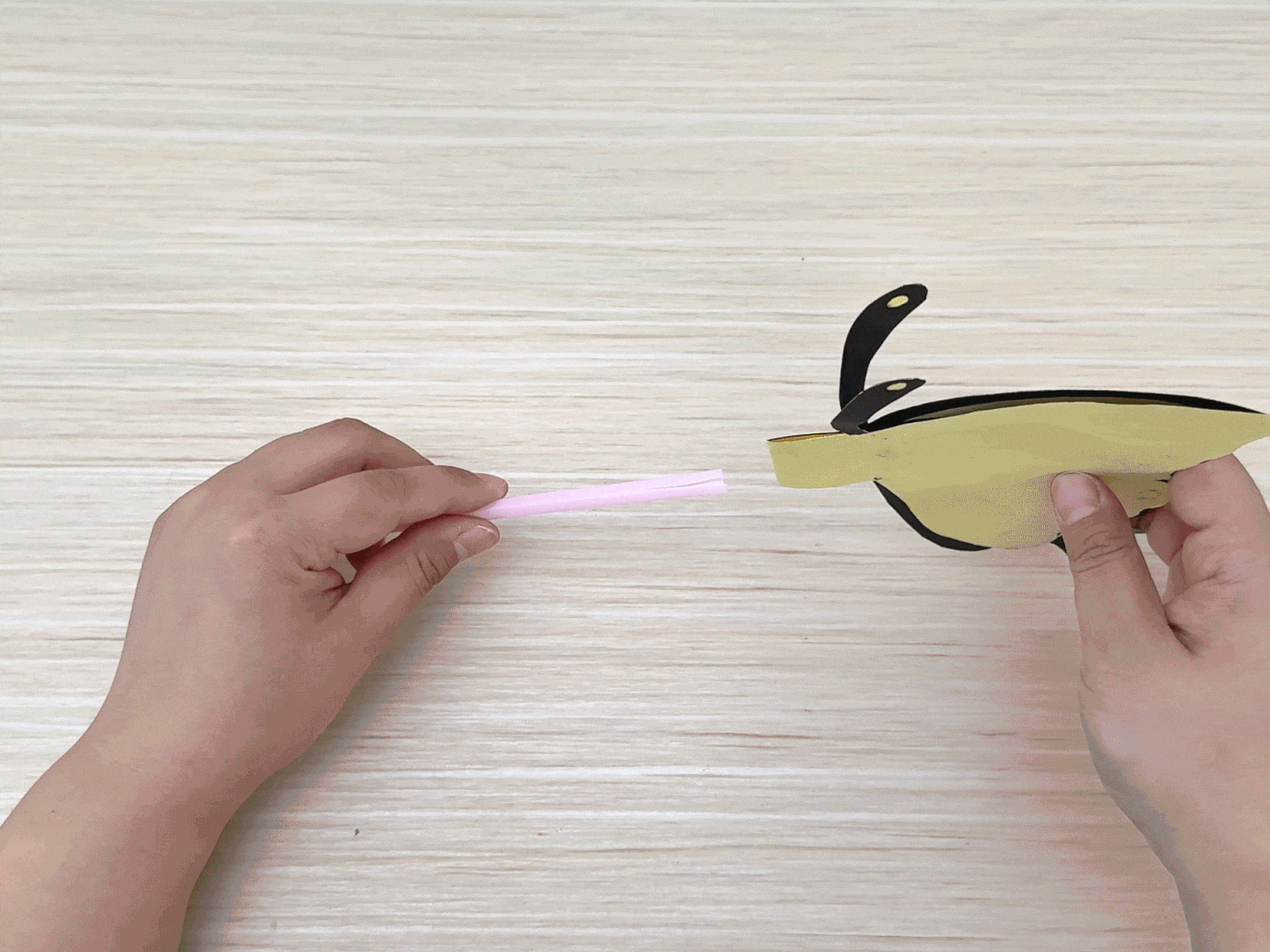 Two hands manipulate elements of a craft project on a light wood background. The left hand holds a short segment of a light pink drinking straw. The right hand holds a partially assembled paper butterfly; its wings are yellow and edged with black, and black antennae protrude upwards. The straw piece is being positioned near the butterfly's body, suggesting the next step in assembling the craft.