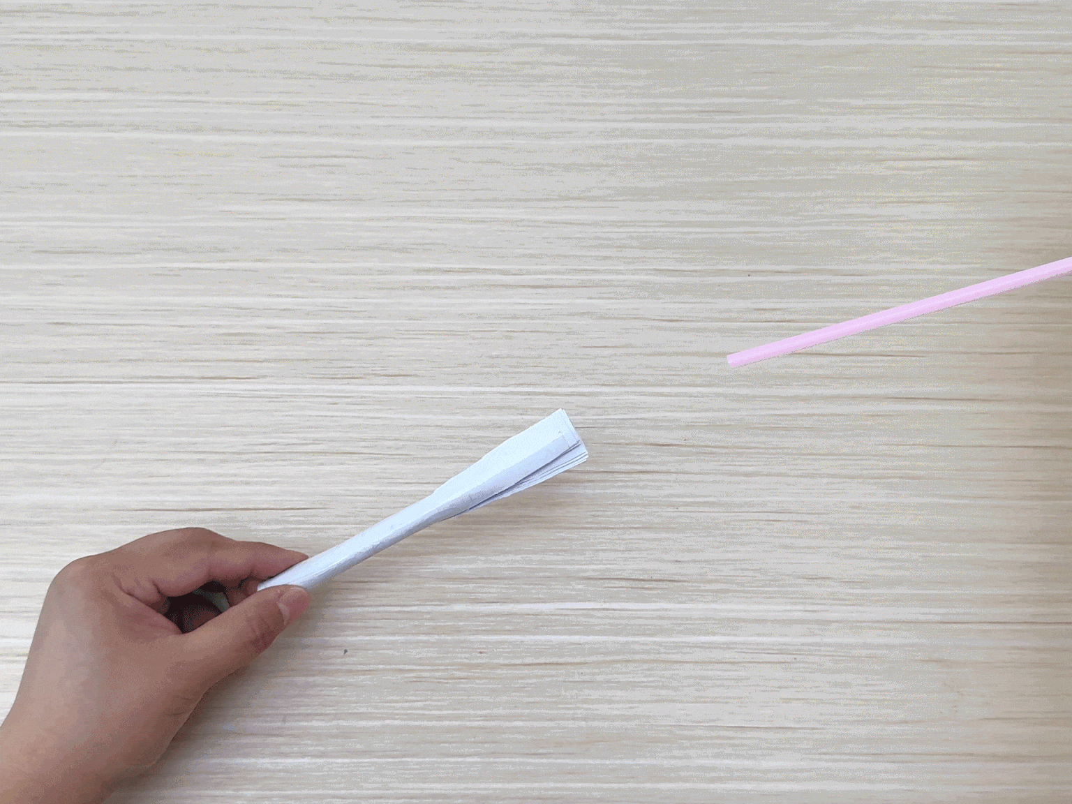 Two hands hold separate parts of a craft project over a light wood-grained surface. The left hand holds a white paper tube, flattened and slightly fanned at one end. The right hand holds a light pink drinking straw. The two items are positioned as if about to be joined together, likely the next step in assembling the craft.