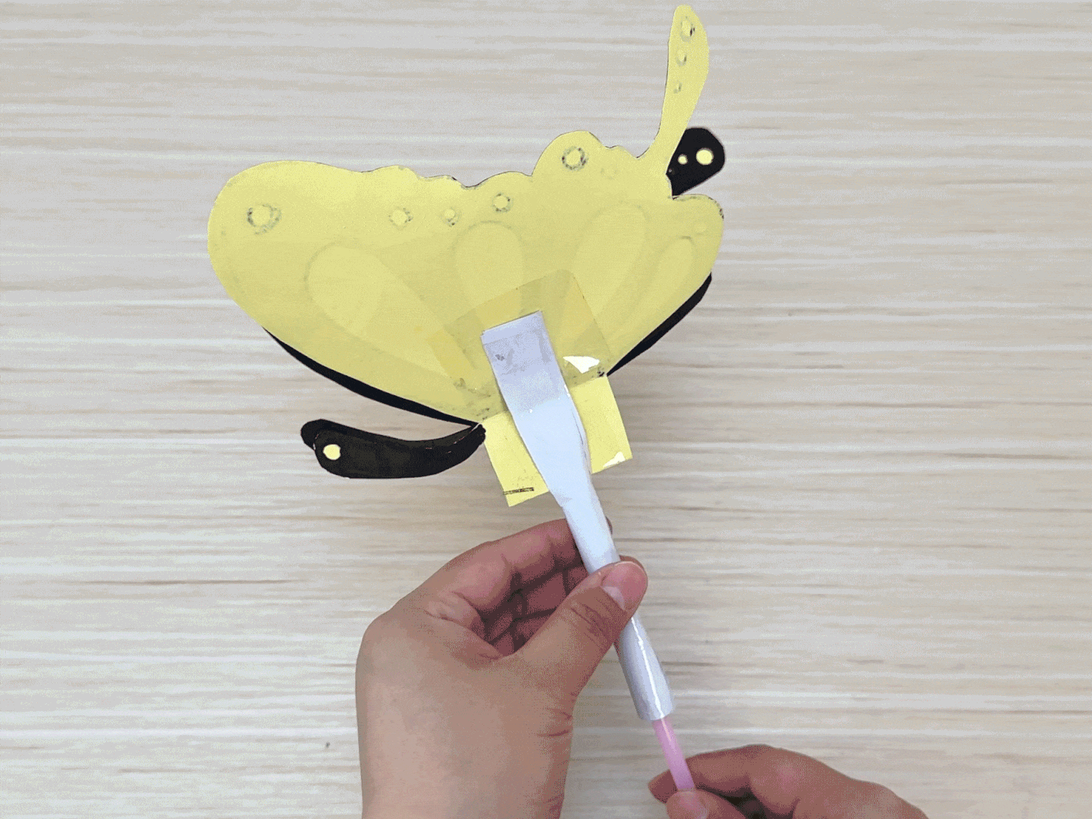Two hands assemble a paper butterfly craft on a light wood background. The butterfly has yellow wings outlined in black, black antennae, and small black details on the wings. A flattened white paper tube, attached to a short pink straw, is being inserted through pre-cut slits in the butterfly's body. Clear tape secures the paper tube to the butterfly's wings. This image captures a stage in the construction of the butterfly, showing the process of attaching the body to the wings.