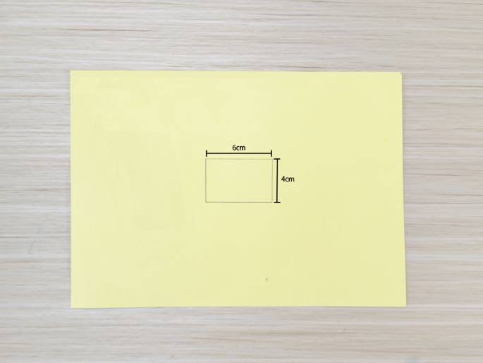 A pale yellow sheet of paper rests on a light wood-grained surface. A smaller rectangle is lightly drawn in the center of the paper, measuring 6cm by 4cm as indicated by lines and labels showing the dimensions. This image likely illustrates a step in a crafting tutorial, showing the planned size and placement of a cut or fold.