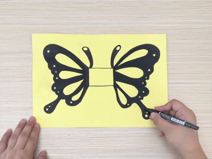 A hand holding a black marker is coloring a butterfly drawing on a piece of pale yellow paper placed on a light wooden surface. The butterfly is drawn in black ink with details on its wings, and a rectangular section in the center of the butterfly's body is left uncolored. The hand is currently coloring the lower portion of the butterfly's right wing. Another hand is partially visible at the left edge, holding the paper steady. This image appears to be from a crafting tutorial or demonstration.