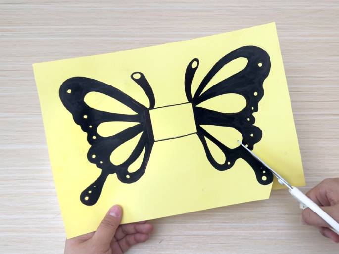 A pair of hands is cutting out a butterfly shape from pale yellow paper with white-handled scissors. The butterfly, drawn in black marker, is positioned centrally on the paper against a light wooden background. The left hand holds the paper while the right hand carefully cuts along the outline of the butterfly's right wing. A rectangular area in the center of the butterfly's body remains uncut. This image is likely part of a crafting tutorial.