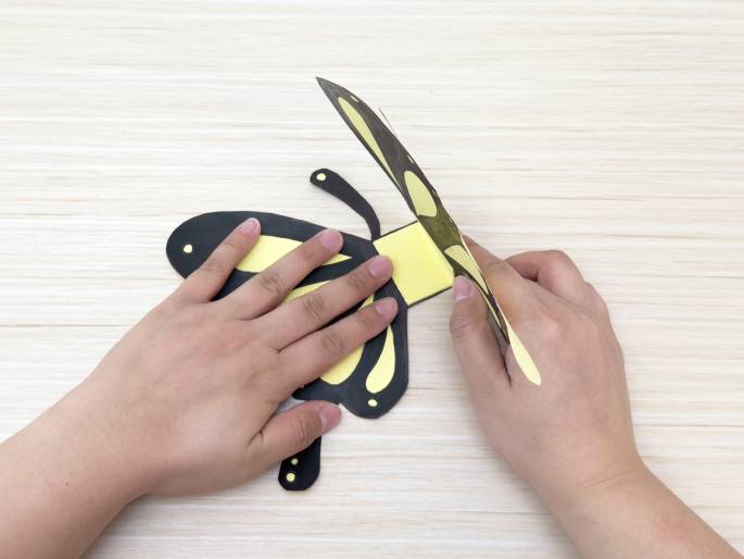 Two hands manipulate a partially constructed paper butterfly against a light wooden background. The butterfly's wings are black with yellow markings and are being folded upward. The left hand holds the lower portion of the butterfly's body, while the right hand adjusts the top wing. The central rectangular section of the butterfly's body is yellow, serving as a hinge for the wings. This image appears to be a step in a paper craft tutorial.