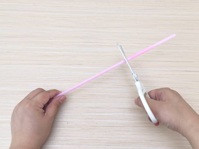 A pair of hands is shown cutting a light pink drinking straw with white-handled scissors. The straw is positioned diagonally across a light wood-grained surface. The left hand holds the straw while the right hand operates the scissors, in the process of cutting the straw. This image likely demonstrates a step in a crafting project.