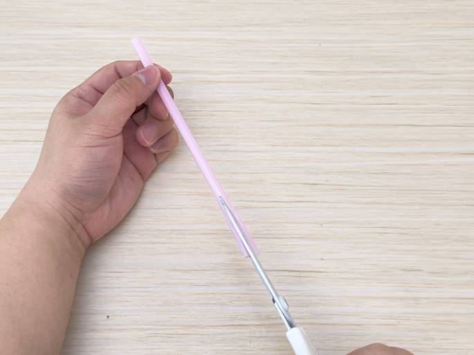 Two hands are cutting a pale pink drinking straw with white-handled scissors on a light-colored wooden surface. The left hand holds the straw vertically while the right hand uses the scissors to make a cut a short distance from the top end. The focus is on the action of cutting the straw. This image likely depicts a step in a craft or DIY project.