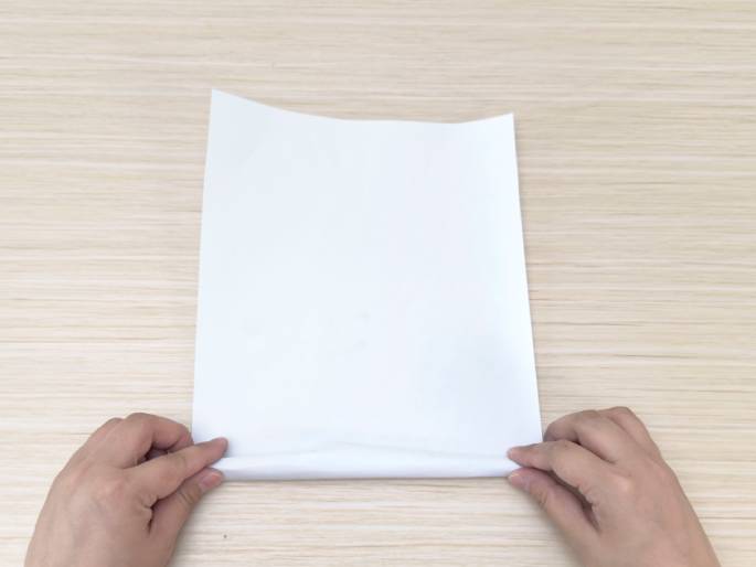 Two hands are beginning to fold a piece of blank white paper on a light wood-grained table. The hands are positioned at the bottom edge of the paper, starting to roll it upwards. The image suggests the first step in a paper folding activity or craft project.