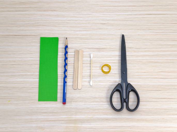 Arranged on a light wood-grained surface are various craft supplies. From left to right: a rectangular piece of bright green paper or fabric, a blue pencil, two small wooden craft sticks, a cotton swab, a small yellow rubber band, and a pair of black scissors. These items appear to be gathered in preparation for a craft project.