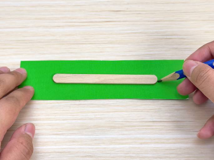A small wooden craft stick rests on a rectangular piece of bright green paper or fabric. A hand holding a blue pencil is marking the green material next to the end of the craft stick, likely measuring or outlining for a craft project. The materials are arranged on a light wood-grain surface.