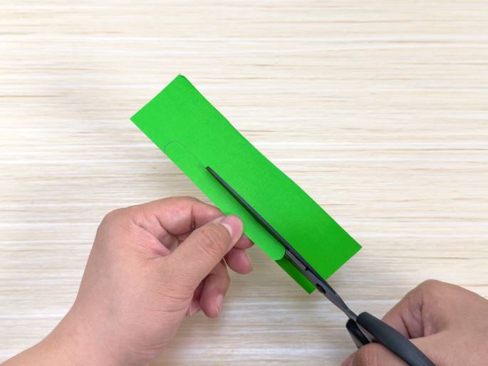 Two hands are shown working with a piece of bright green paper or fabric on a light wood-grain surface. One hand holds the green material while the other uses black scissors to cut a rounded shape along one edge. This image likely depicts a step in a craft project.
