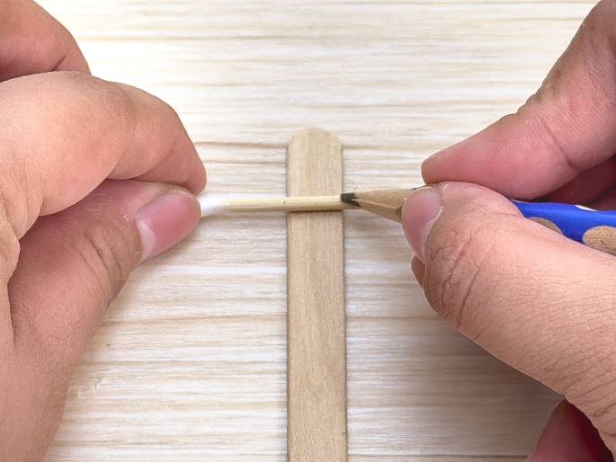 Against a light wood-grain background, two hands are using a cotton swab and a blue pencil to mark a wooden craft stick. One hand holds the cotton swab across the craft stick, while the other hand uses the pencil to mark the stick along the edge of the swab. This suggests measuring or preparing the stick for some kind of craft or construction activity.