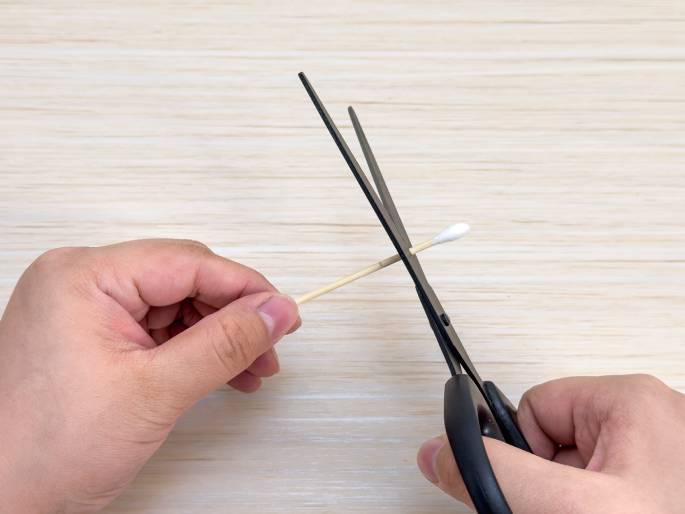 Two hands are shown cutting a cotton swab with a pair of black scissors. One hand holds the cotton swab against a light wood-grain background, while the other hand uses the scissors to cut the swab's wooden stick.