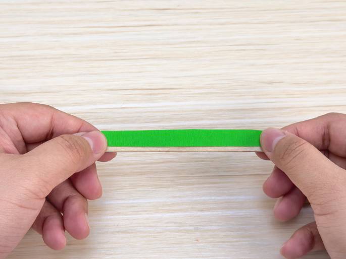 Two hands hold a thin wooden craft stick covered with a strip of bright green paper or tape. The stick is held horizontally against a light wood-grain background. The image likely depicts a stage in a craft project.