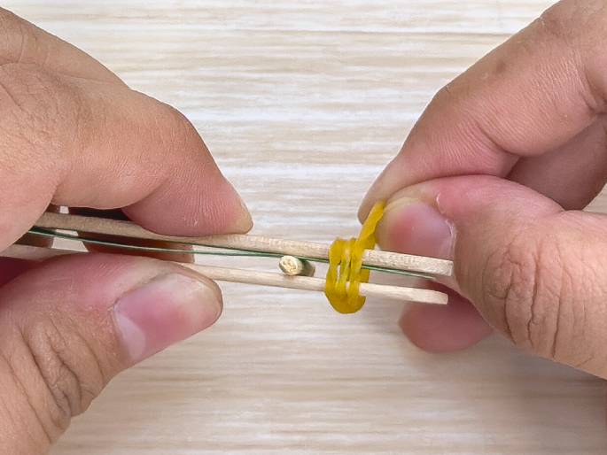 Two hands are manipulating small wooden craft sticks and a yellow rubber band against a light wood-grain background. A small, cylindrical object is held between the sticks, and the rubber band is wrapped around them, seemingly securing the object in place. This image likely depicts a step in constructing a small craft or toy.