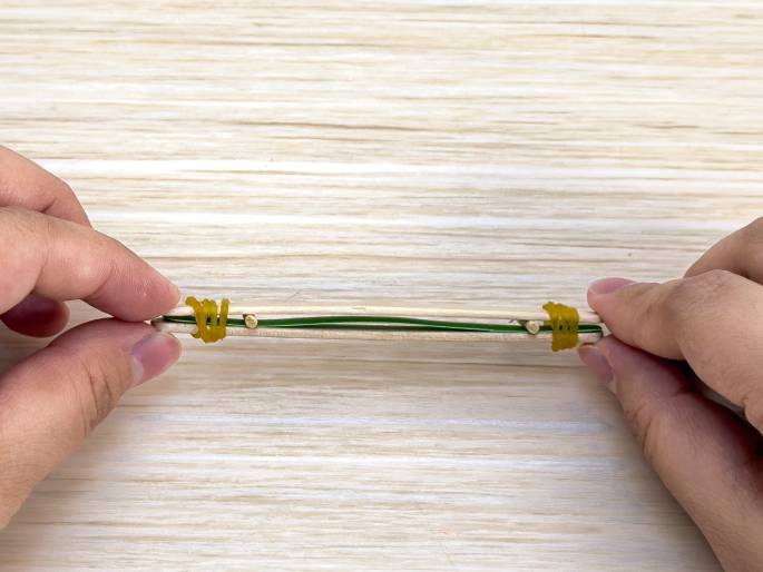 Two hands hold a small, simple construction made of wooden craft sticks, small wooden dowels, and yellow rubber bands. A thin green wire or string is stretched between the dowels, which are attached to the craft sticks. The rubber bands appear to be holding the structure together. The background is a light wood-grain surface. This close-up image likely shows a step in a craft project, possibly building a miniature catapult or similar device.