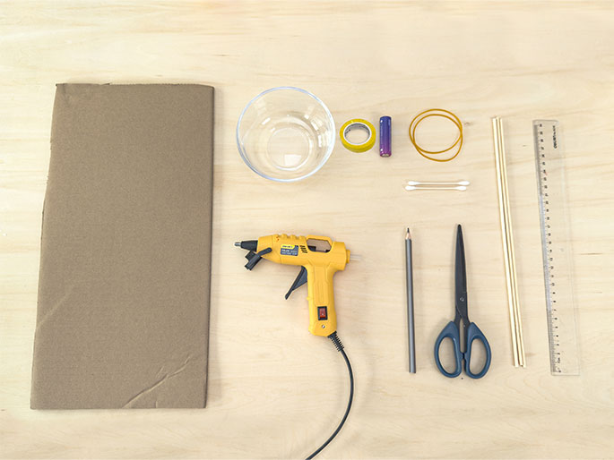 A collection of crafting materials is laid out on a light woodgrain work surface, viewed from above. The items include a rectangular piece of brown cardboard, a clear glass bowl, a roll of yellow tape, a small purple battery, a rubber band, two cotton swabs, a yellow hot glue gun with its cord visible, a gray pencil, a pair of blue-handled scissors, two thin wooden dowels, and a clear plastic ruler. The arrangement suggests the preparation for a DIY or crafting project.