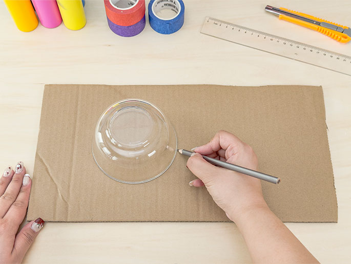 On a light wooden table, a piece of brown cardboard lies flat, with a clear glass bowl placed on top. A hand holding a gray pencil is tracing the rim of the bowl onto the cardboard. Additional crafting supplies, including colorful rolls of tape, a ruler, and a utility knife, are visible in the background, suggesting this tracing is a step in a larger crafting process. The hand tracing has fingernails decorated with a design.
