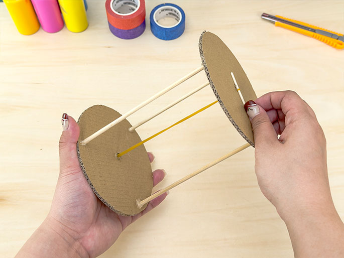 Two hands hold a cylindrical structure made of two cardboard circles connected by three wooden dowels. A yellow rubber band is stretched between the two cardboard circles, passing through central holes in each. The hands, with fingernails decorated with a floral design, appear to be adjusting the rubber band. In the background, on a light wood surface, are various craft supplies, including paint bottles and rolls of colorful tape, along with a utility knife.