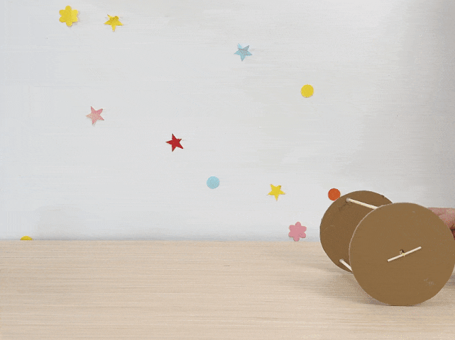 A small toy car made of cardboard circles for wheels, wooden skewers as axles, and a yellow clay or playdough center rolls back and forth on a light wooden surface. The backdrop is a white wall adorned with colorful paper stars, dots, and flowers. A hand briefly enters the frame to push the toy. This short video demonstrates a simple craft activity, likely for children.